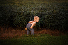 Chambray blue long overalls on a little boy model showcasing the back while playing in the summer - Handmade from repurposed linen fabric, these gender-neutral overalls are ideal for passing down. The breathable fabric ensures comfort, while stylish pleats add elegance. Durable leather-looking straps enhance visual appeal, and adjustable straps in two lengths provide a perfect fit. Unfoldable leg cuffs designed to grow with your little one make these overalls a practical and sustainable choice for active ki