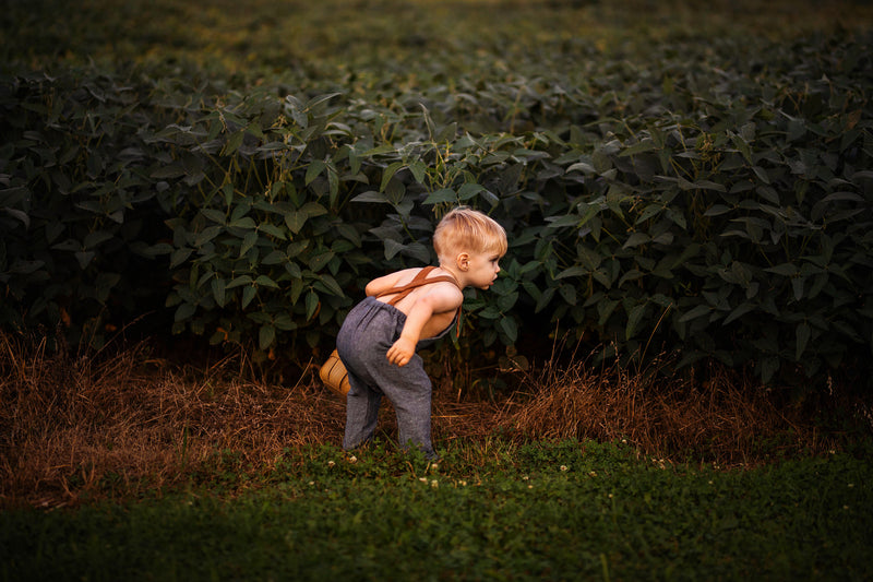 Chambray blue long overalls on a little boy model showcasing the back while playing in the summer - Handmade from repurposed linen fabric, these gender-neutral overalls are ideal for passing down. The breathable fabric ensures comfort, while stylish pleats add elegance. Durable leather-looking straps enhance visual appeal, and adjustable straps in two lengths provide a perfect fit. Unfoldable leg cuffs designed to grow with your little one make these overalls a practical and sustainable choice for active ki