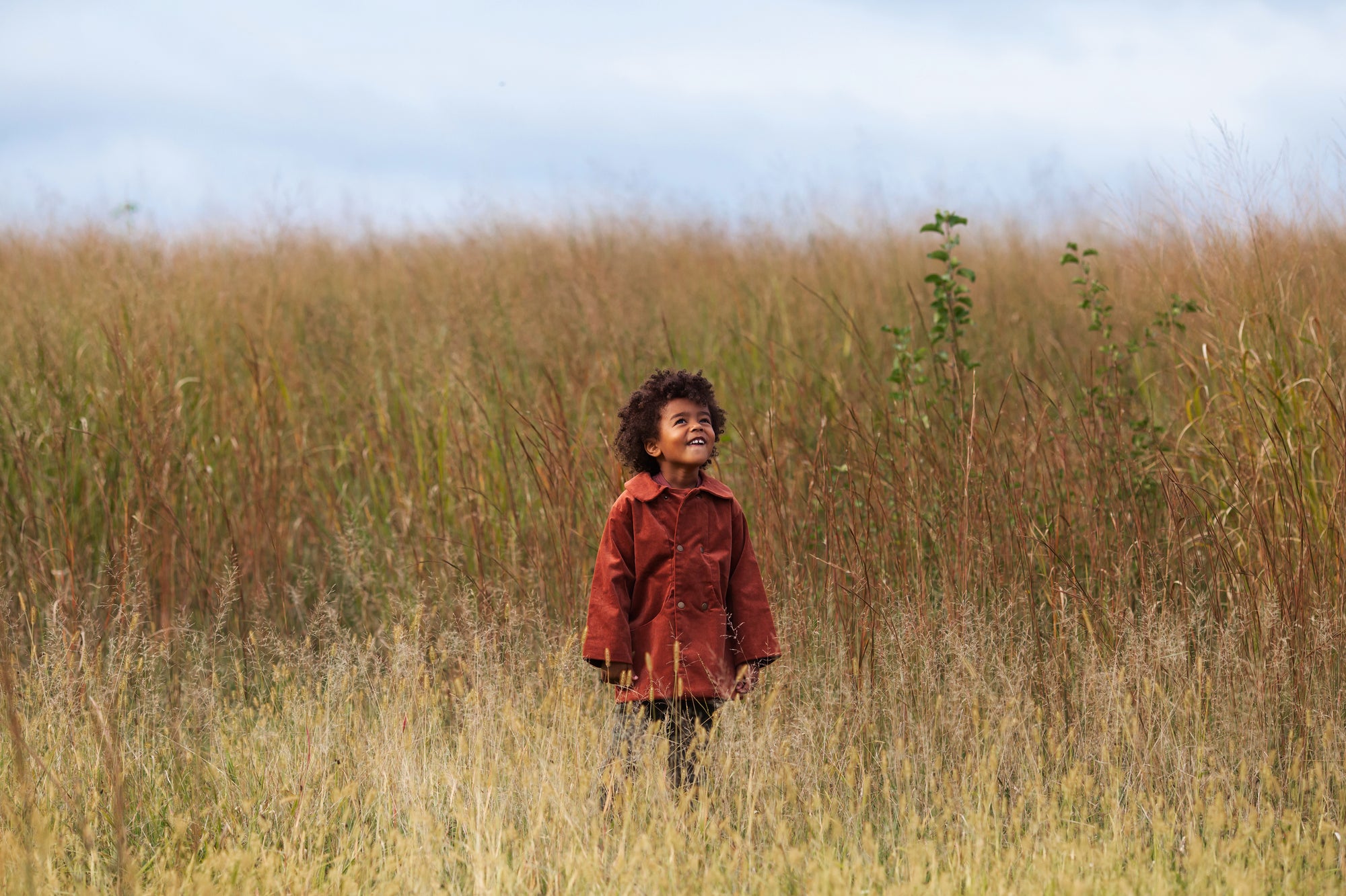 Child in a red jacket standing in tall grass with a clear sky.
