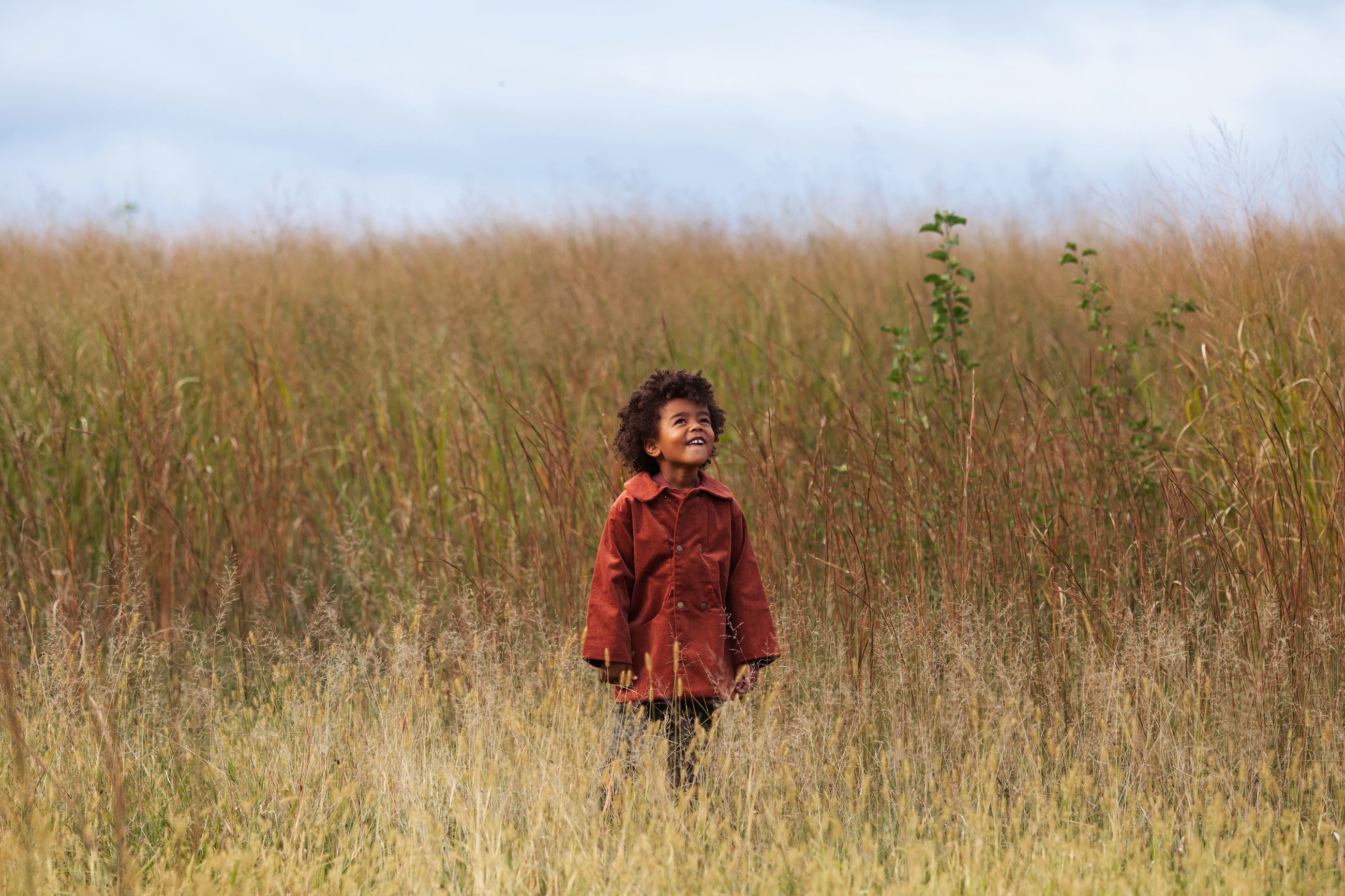 Child in a red jacket standing in tall grass with a clear sky.