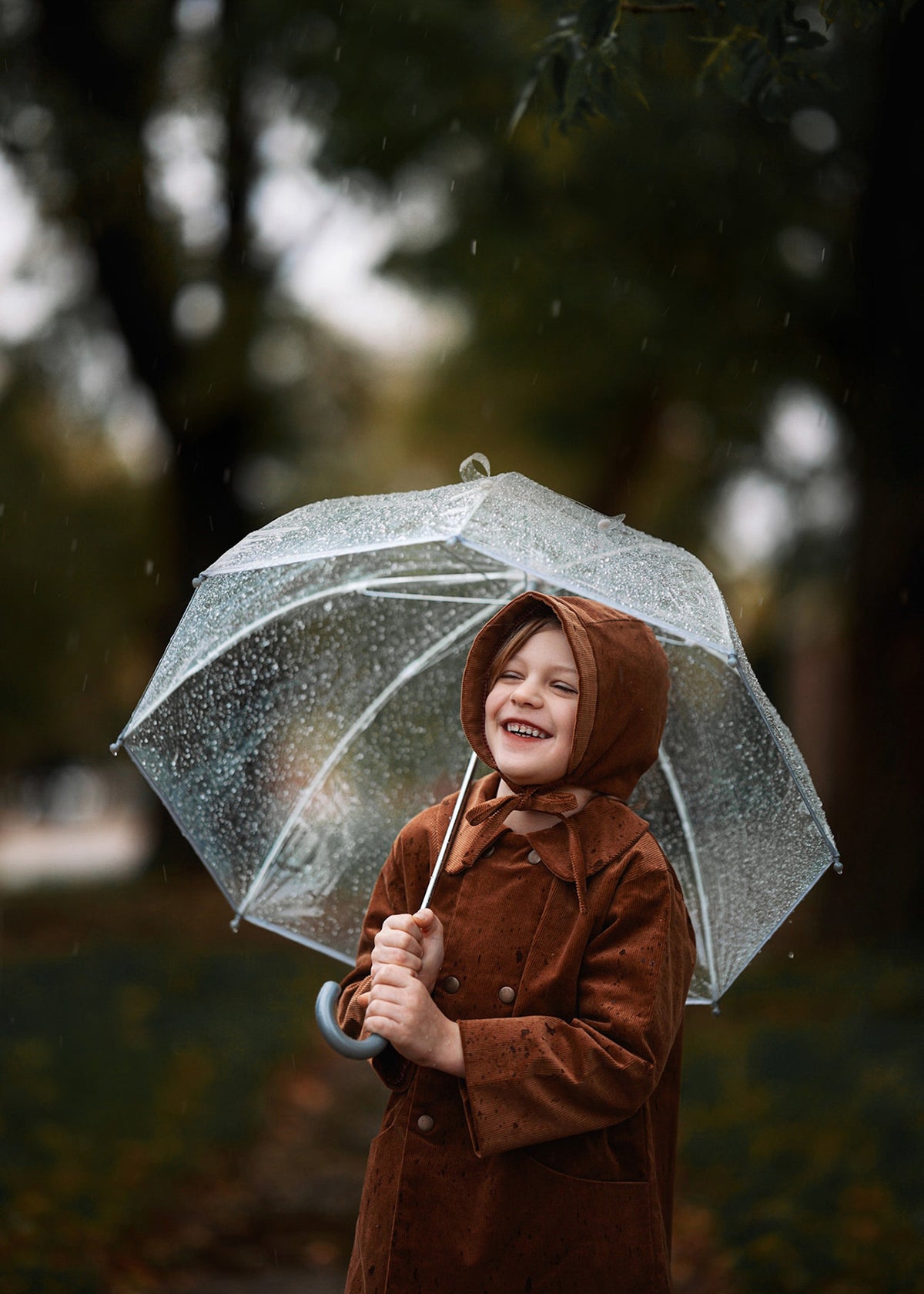 Child in a brown coat and hood holding a transparent umbrella in a rainy setting