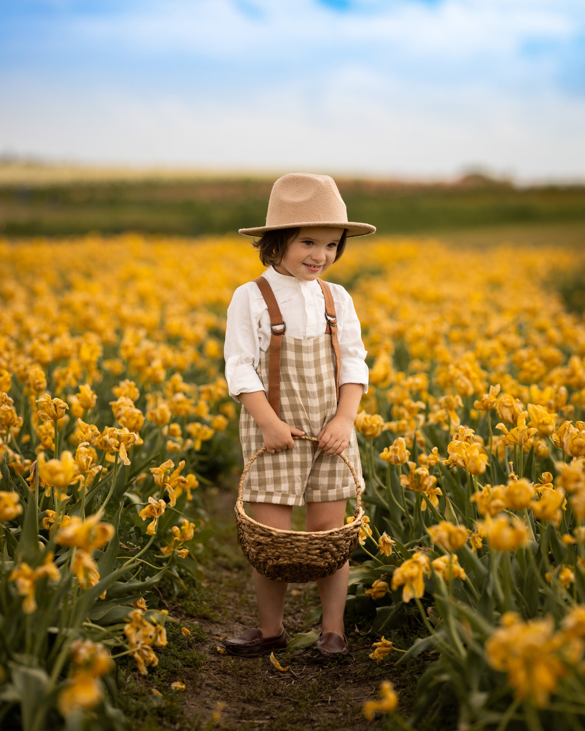 Child in a hat and checkered dress standing in a field of yellow flowers holding a basket.