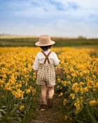 Little boy seeing from the back in a beige gingham romper surrounded by yellow flowers in a field.