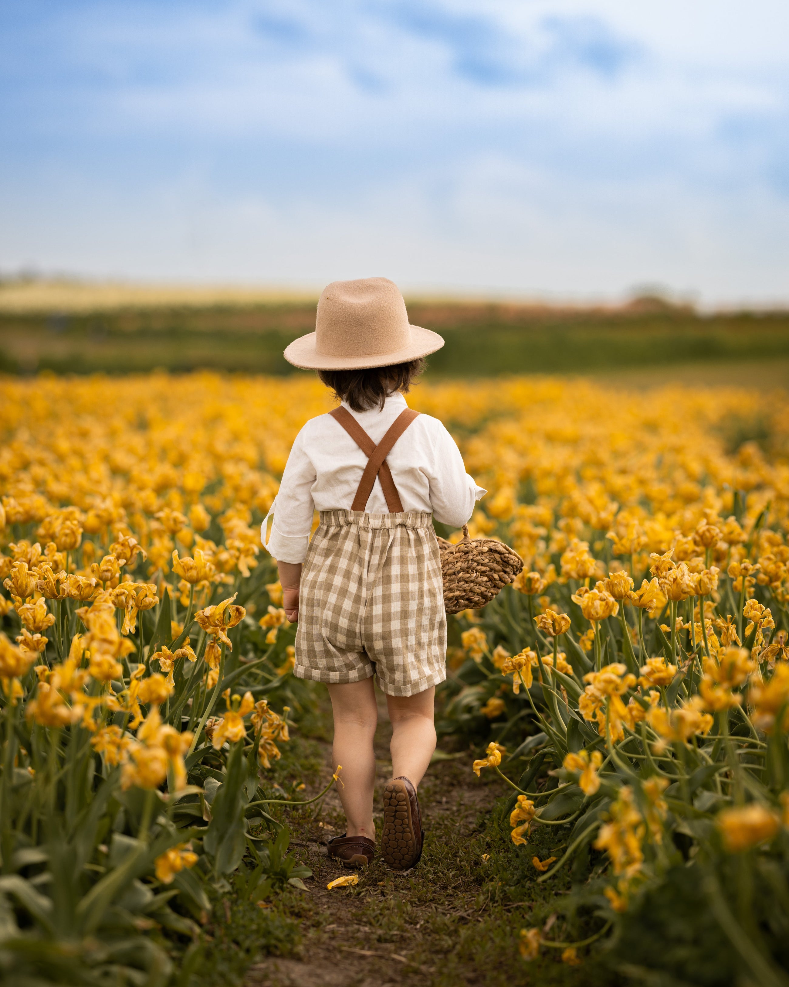 Little boy seeing from the back in a beige gingham romper surrounded by yellow flowers in a field.
