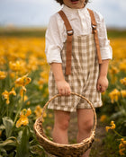 Little boy in a beige gingham romper surrounded by yellow flowers in a field.
