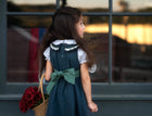 A young girl with long dark hair is shown from behind, wearing a dark teal pinafore dress with a white ruffled collar and a large teal bow at the back. She is holding a wicker basket filled with red flowers, standing in front of a window with a warm sunset reflection.