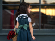 A young girl with long dark hair is shown from behind, wearing a dark teal pinafore dress with a white ruffled collar and a large teal bow at the back. She is holding a wicker basket filled with red flowers, standing in front of a window with a warm sunset reflection.