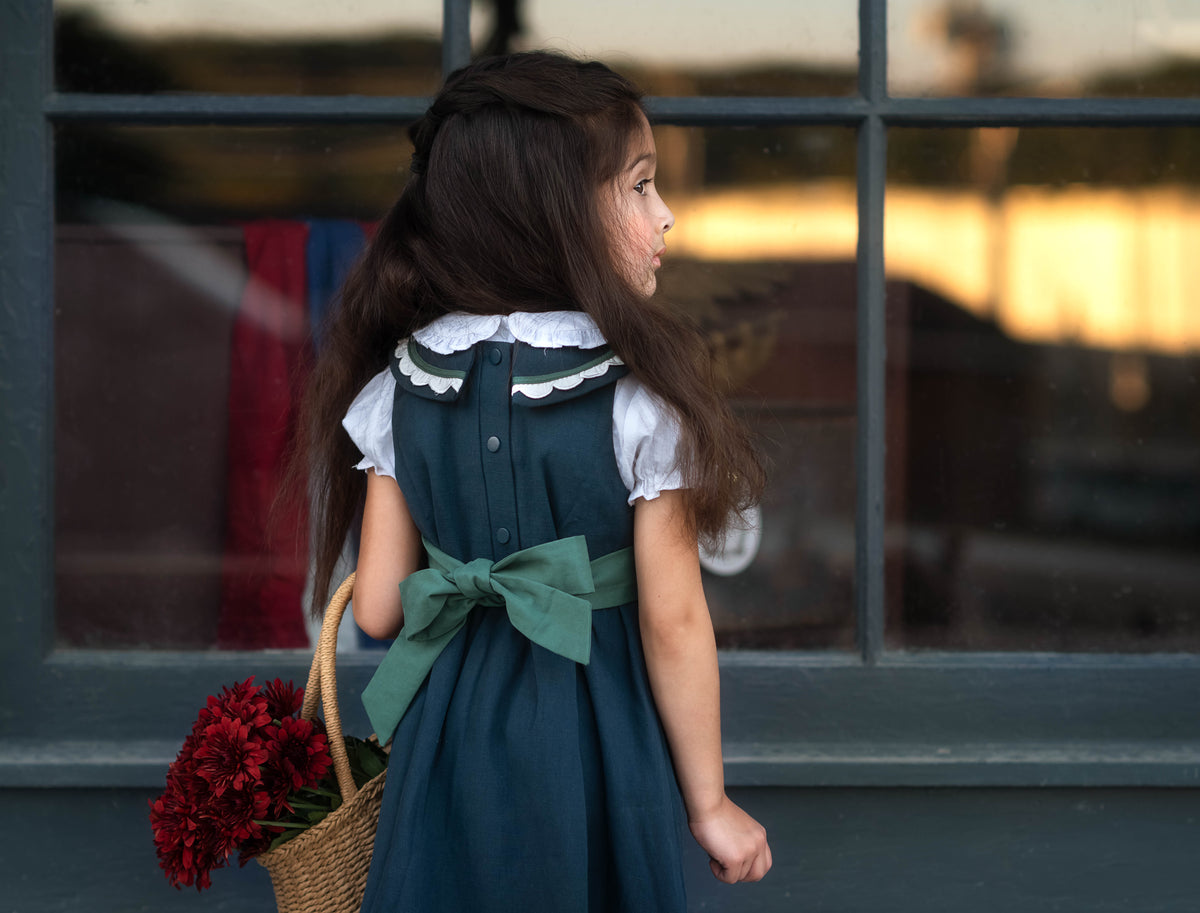 A young girl with long dark hair is shown from behind, wearing a dark teal pinafore dress with a white ruffled collar and a large teal bow at the back. She is holding a wicker basket filled with red flowers, standing in front of a window with a warm sunset reflection.
