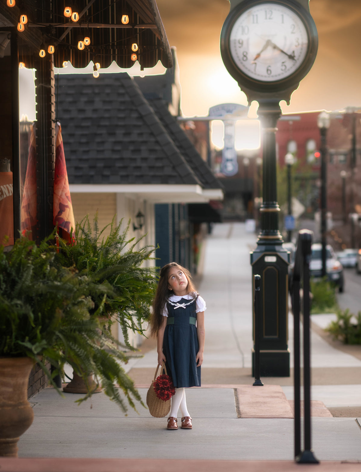 A young girl stands on a city sidewalk wearing a navy blue and white sailor-style dress, holding a basket with red flowers, with a large black street clock and storefronts in the background.