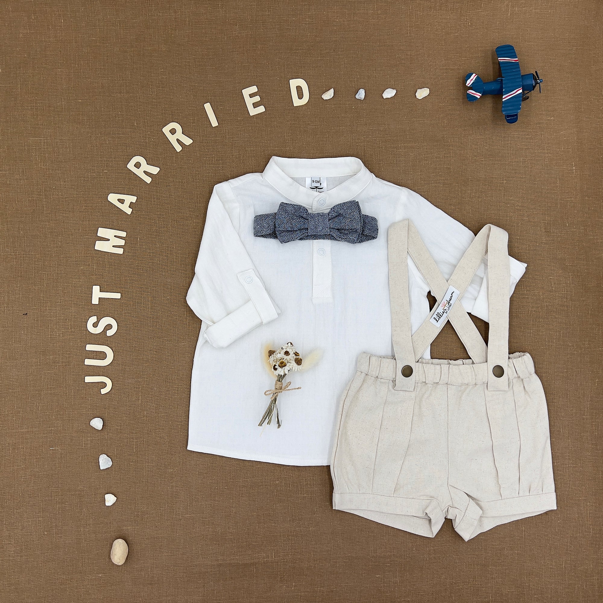 A baby boy's two-piece clothing set featuring a white long-sleeve shirt with a gray patterned bow tie and cream-colored linen suspenders, styled on a brown textured background with decorative props and 'JUST MARRIED' lettering.