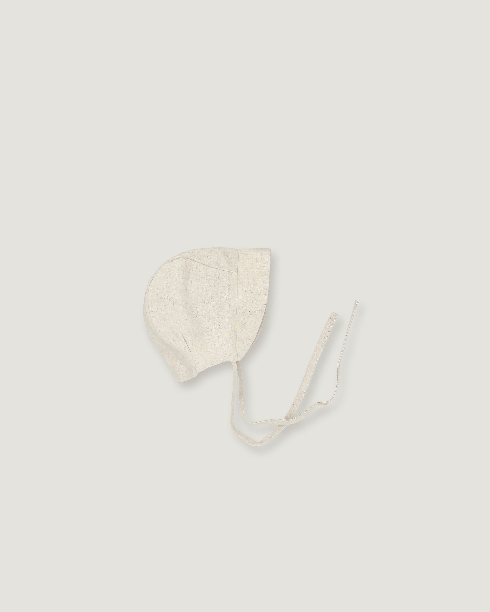 A light cream-colored linen bonnet with long ties, photographed flat on a matching light gray background.