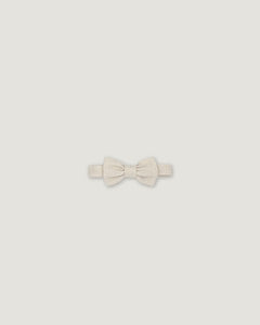 A small bowtie in a textured cream or off-white fabric, centered on a solid light gray background.