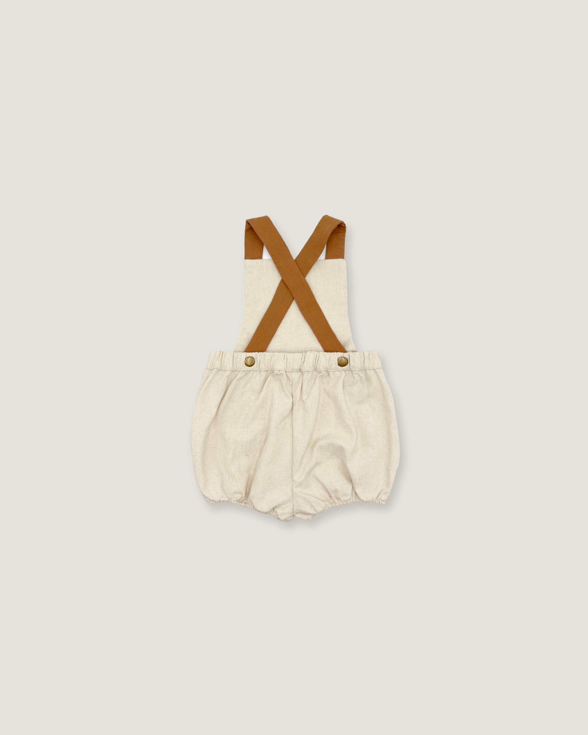A baby romper with cream-colored fabric and brown suspenders, shown flat on a light gray background.