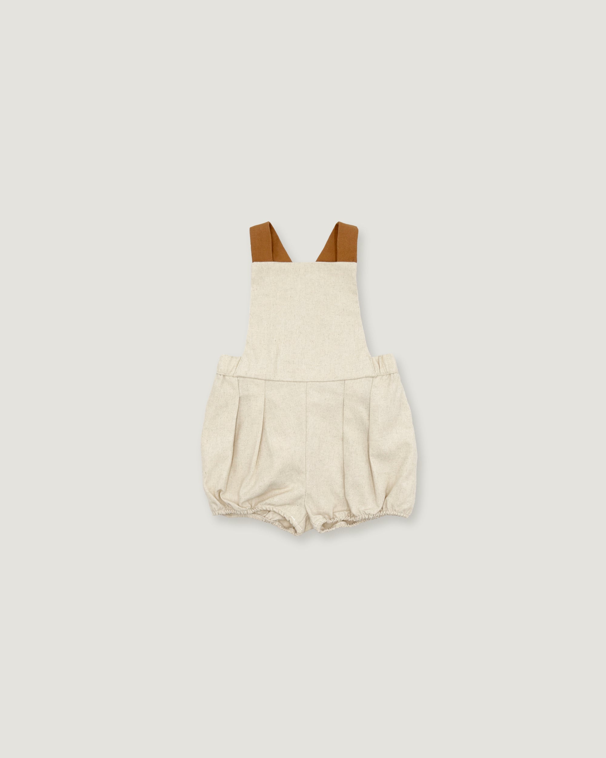 A baby romper in a textured cream linen-like fabric, featuring brown shoulder straps, displayed flat against a solid light gray background.