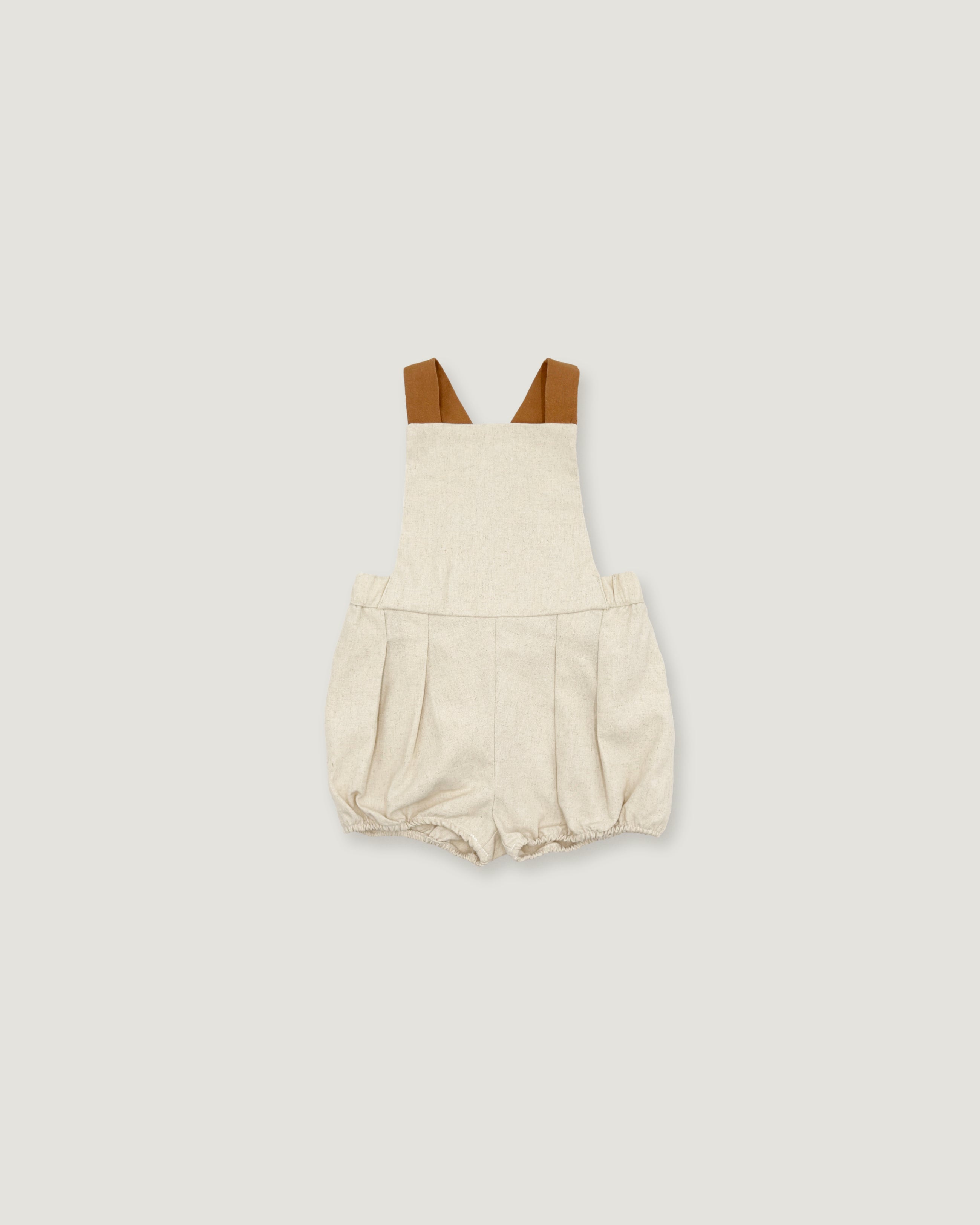 A baby romper in a textured cream linen-like fabric, featuring brown shoulder straps, displayed flat against a solid light gray background.