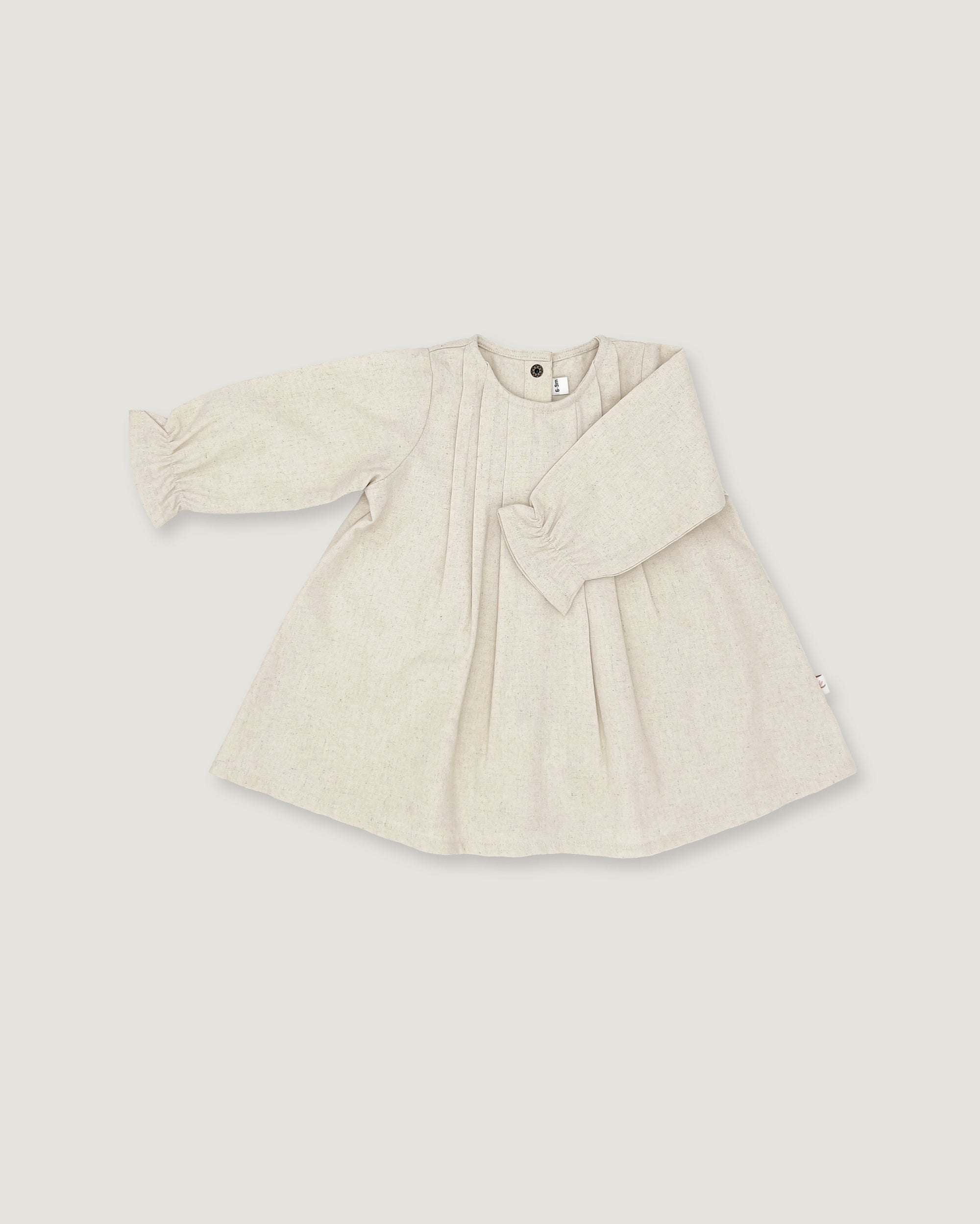A light beige linen long-sleeve dress for toddlers, featuring a gathered waist with a bow detail, pleated front, and elasticized cuffs, shown on a neutral light gray background.
