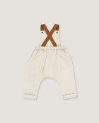 A pair of baby bib pants in a light cream linen-like fabric, featuring brown faux-leather suspenders that cross over the chest. The pants have an elastic waistband and rolled cuffs, shown against a solid light gray background.