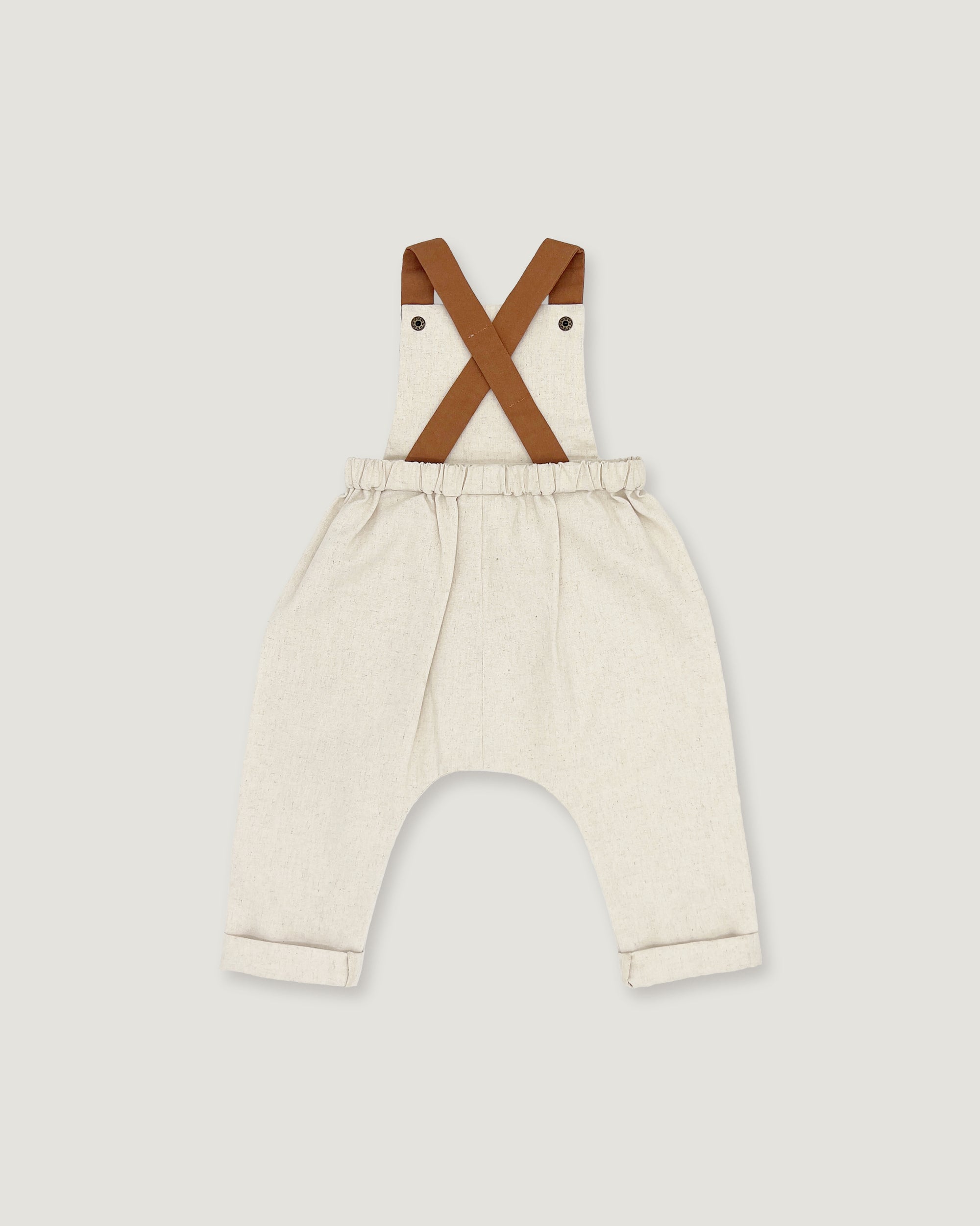 A pair of baby bib pants in a light cream linen-like fabric, featuring brown faux-leather suspenders that cross over the chest. The pants have an elastic waistband and rolled cuffs, shown against a solid light gray background.