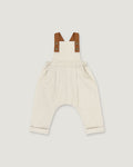 A baby romper in off-white linen with brown leather-like shoulder straps, shown flat on a light gray background.