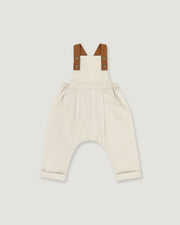 A baby romper in off-white linen with brown leather-like shoulder straps, shown flat on a light gray background.