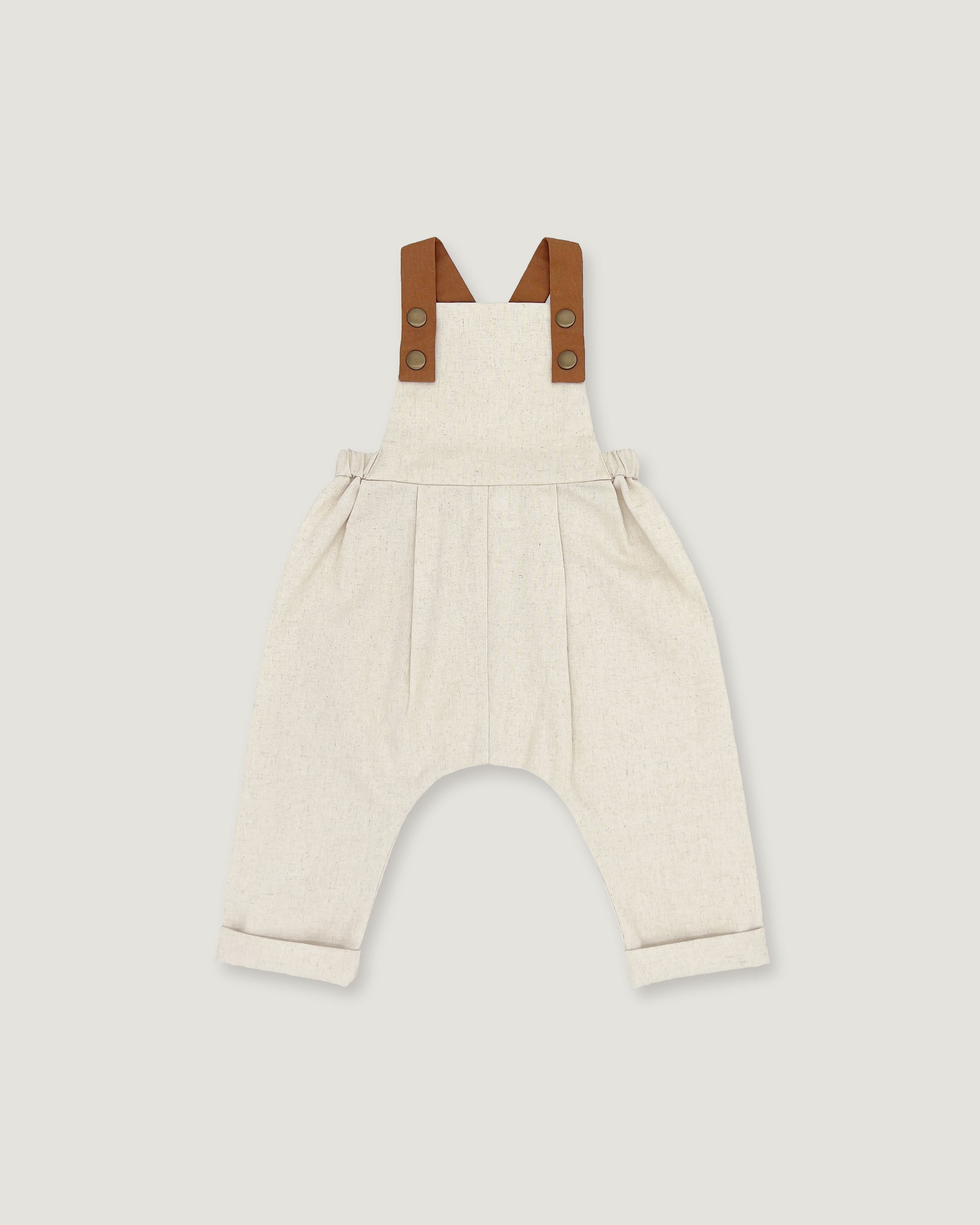 A baby romper in off-white linen with brown leather-like shoulder straps, shown flat on a light gray background.