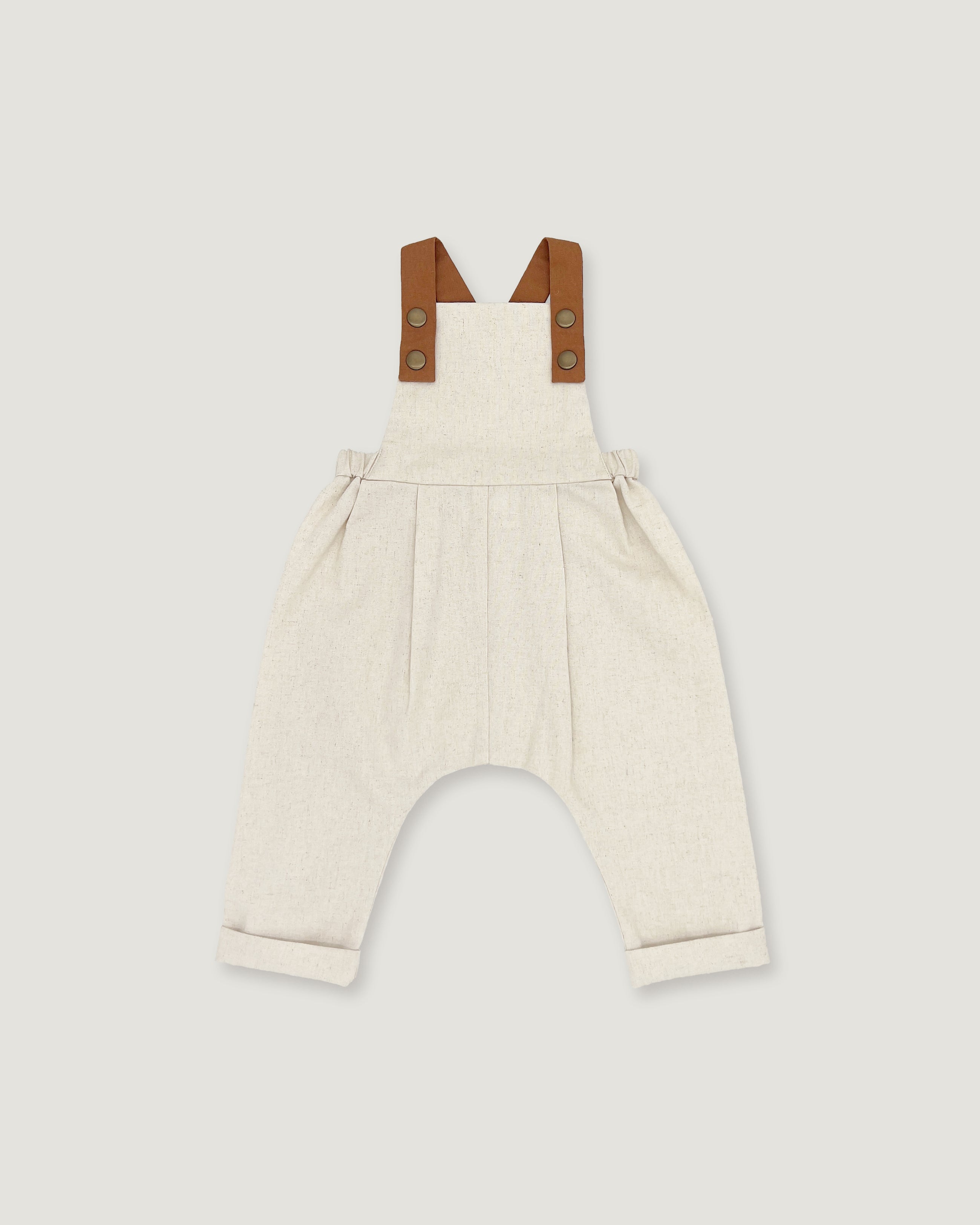 A baby romper in off-white linen with brown leather-like shoulder straps, shown flat on a light gray background.