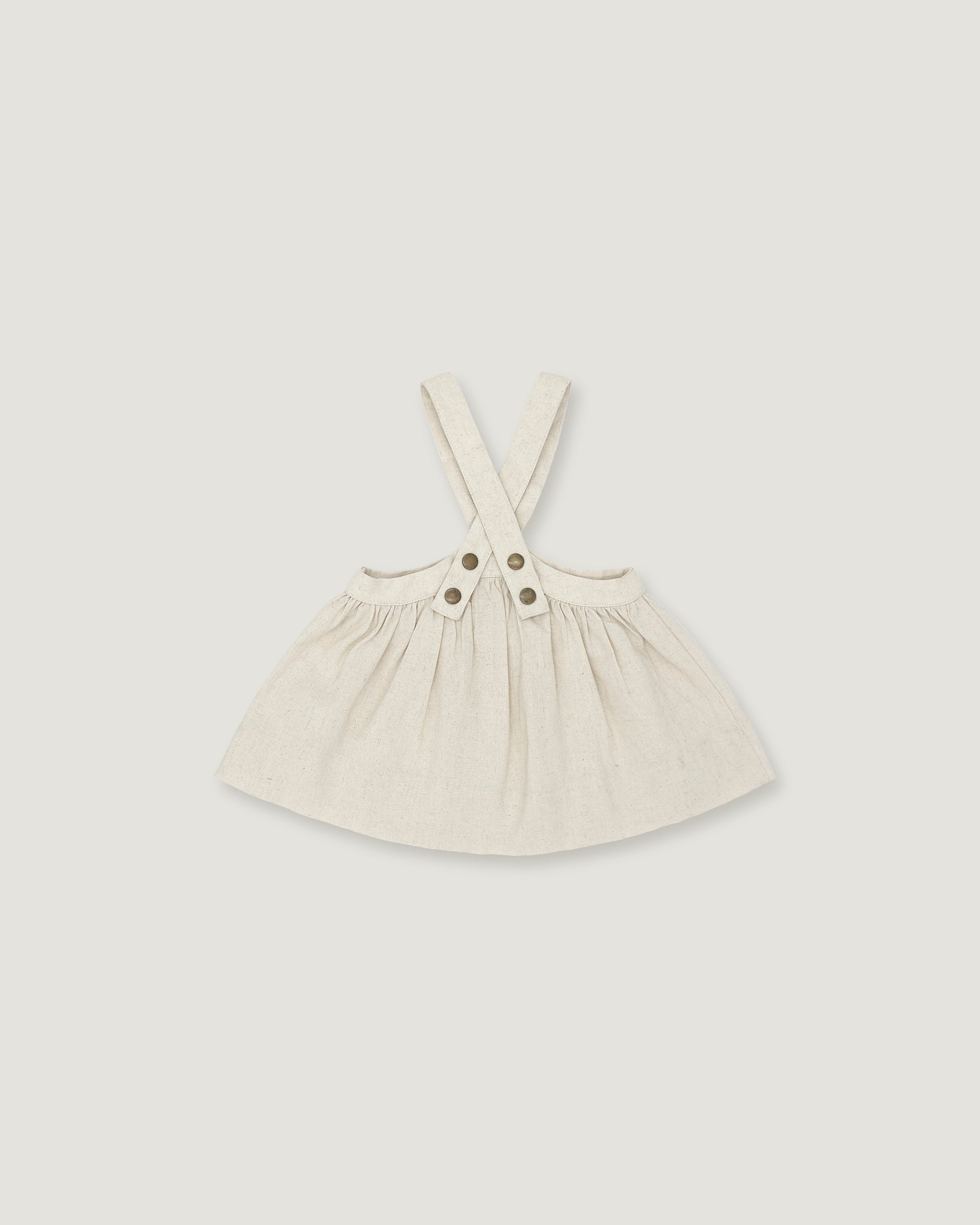A light cream-colored linen pinafore dress for toddlers, featuring adjustable shoulder straps with two decorative buttons at the center, displayed on a neutral light gray background.