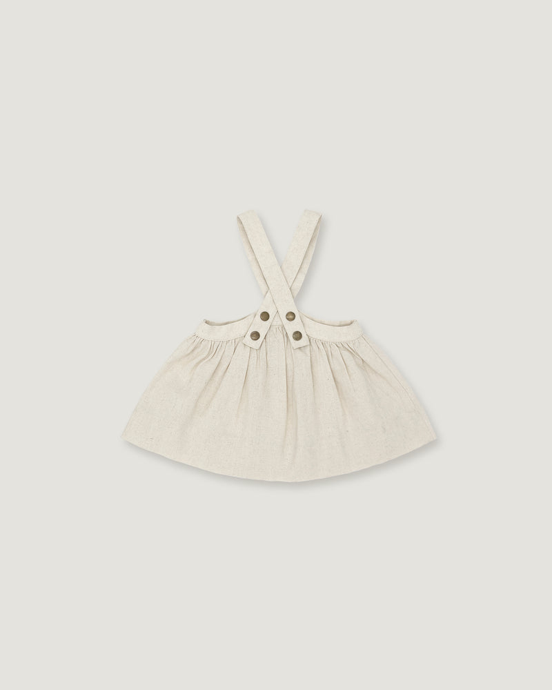 A light cream-colored linen pinafore dress for toddlers, featuring adjustable shoulder straps with two decorative buttons at the center, displayed on a neutral light gray background.