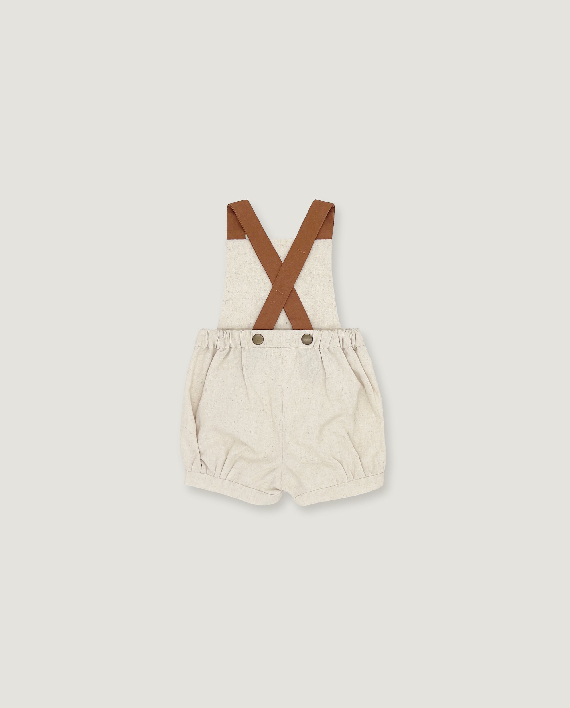 A baby romper with cream-colored fabric and brown suspenders, laid flat against a light gray background.