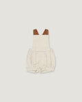 A baby romper in a light oatmeal-beige linen-like fabric, featuring brown faux-leather shoulder straps, shown flat on a light gray background.