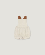 A baby romper in a light oatmeal-beige linen-like fabric, featuring brown faux-leather shoulder straps, shown flat on a light gray background.