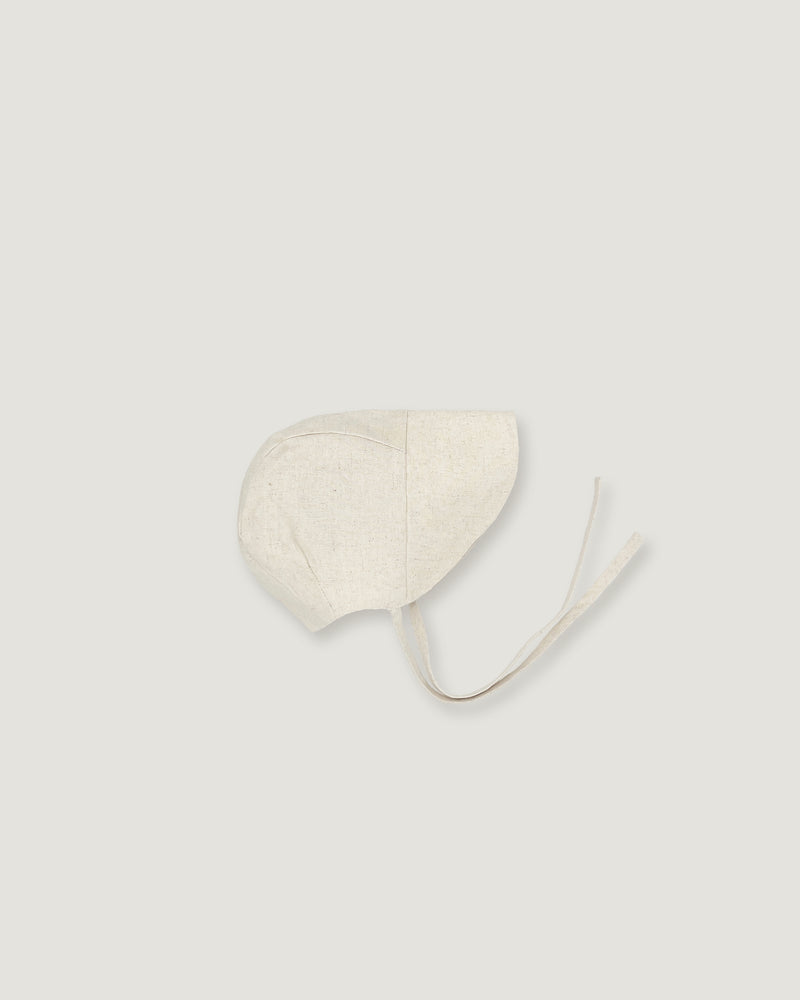 A light cream-colored fabric bonnet with long ties, photographed flat against a solid, very light gray background.