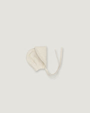 A cream-colored cotton bonnet with tie strings, photographed flat against a solid light gray background.