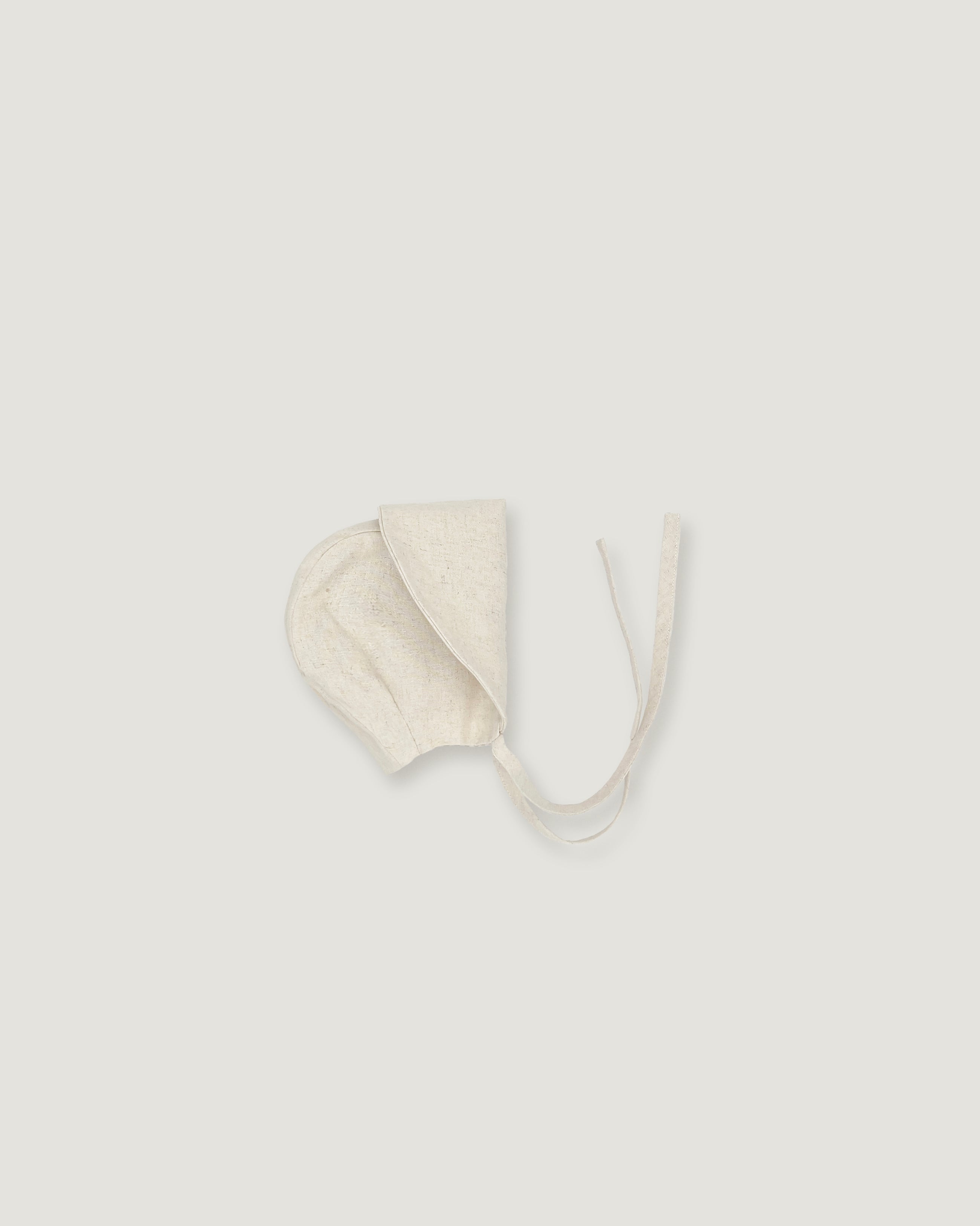 A cream-colored cotton bonnet with tie strings, photographed flat against a solid light gray background.