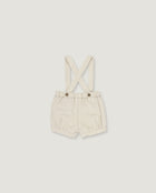 A pair of cream-colored linen baby shorts with adjustable shoulder straps and two metal snap buttons, displayed flat against a light gray background.