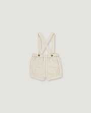 A pair of cream-colored linen baby shorts with adjustable shoulder straps and two metal snap buttons, displayed flat against a light gray background.