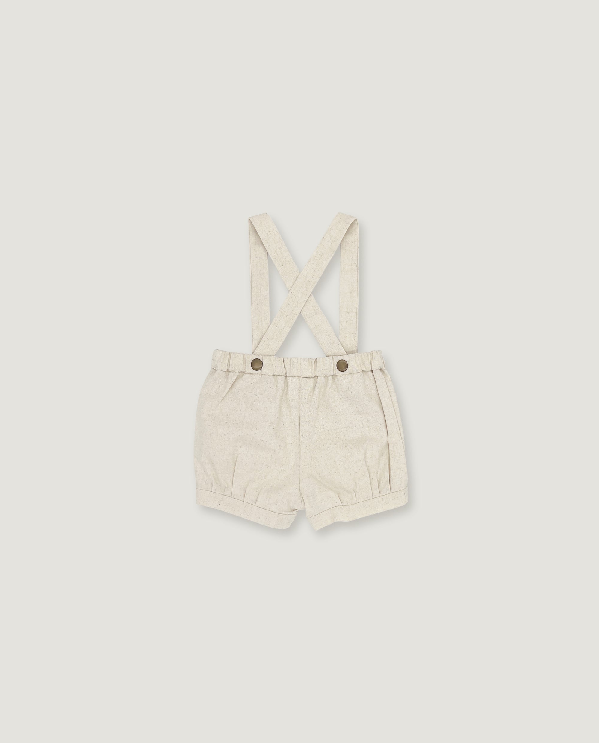 A pair of cream-colored linen baby shorts with adjustable shoulder straps and two metal snap buttons, displayed flat against a light gray background.