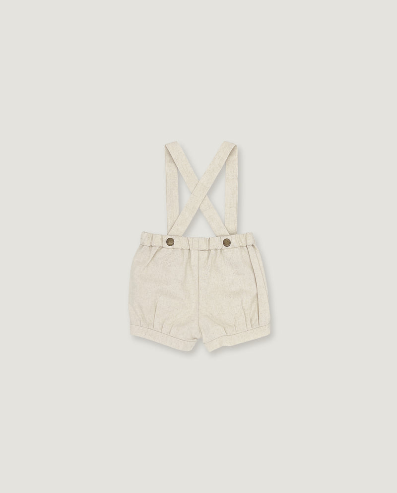 A pair of cream-colored linen baby shorts with adjustable shoulder straps and two metal snap buttons, displayed flat against a light gray background.
