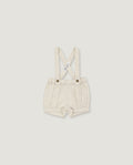 A pair of baby boy's cream-colored linen suspenders shorts with adjustable shoulder straps and metal snap buttons, shown flat on a light gray background.