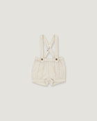 A pair of baby boy's cream-colored linen suspenders shorts with adjustable shoulder straps and metal snap buttons, shown flat on a light gray background.