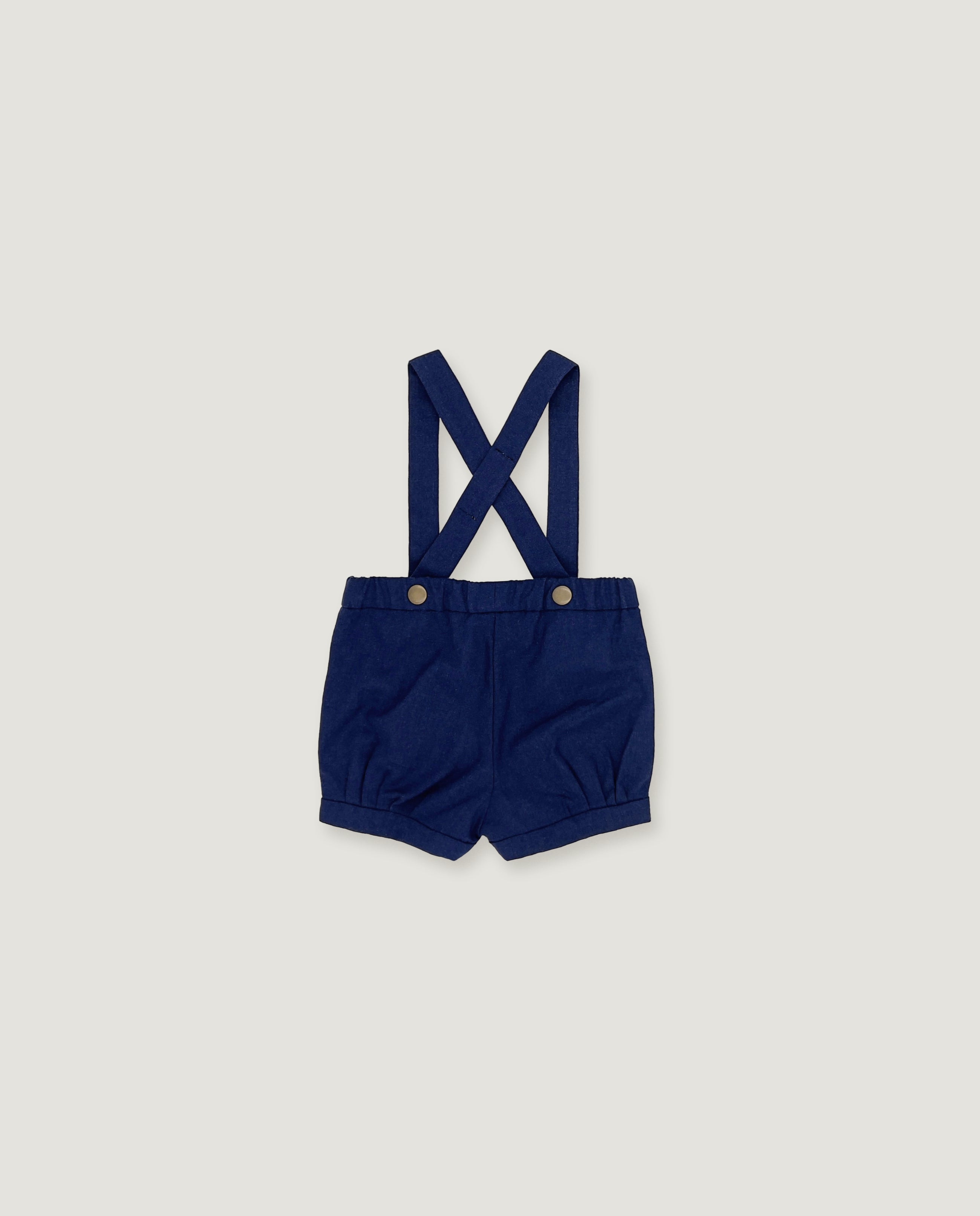A pair of navy blue baby boy shorts with adjustable shoulder straps and metal snap buttons, laid flat on a light gray background.