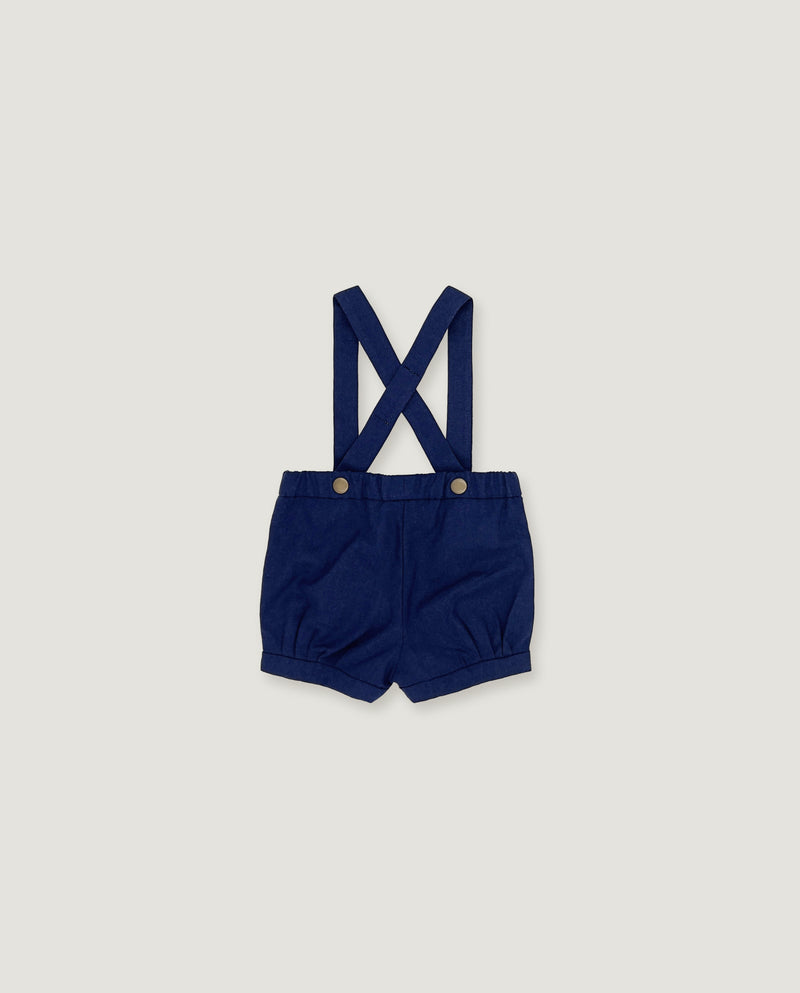 A pair of navy blue baby boy shorts with adjustable shoulder straps and metal snap buttons, laid flat on a light gray background.
