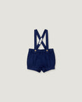 A pair of navy blue baby boy shorts with adjustable suspenders, featuring a white fabric brand tag and gold-tone snap buttons, displayed flat against a light gray background.