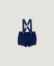 A pair of navy blue baby boy shorts with adjustable suspenders, featuring a white fabric brand tag and gold-tone snap buttons, displayed flat against a light gray background.