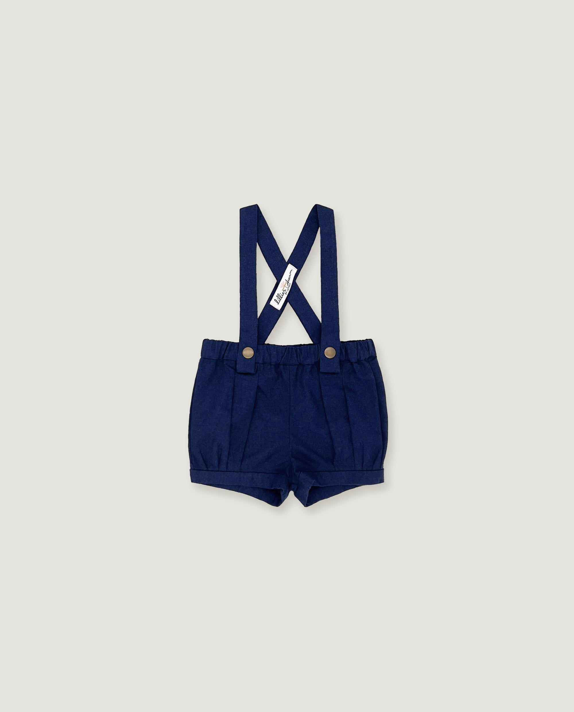 A pair of navy blue baby boy shorts with adjustable suspenders, featuring a white fabric brand tag and gold-tone snap buttons, displayed flat against a light gray background.