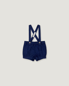 A pair of navy blue baby boy shorts with adjustable suspenders, featuring a white fabric brand tag and gold-tone snap buttons, displayed flat against a light gray background.