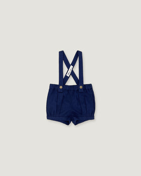 A pair of navy blue baby boy shorts with adjustable suspenders, featuring a white fabric brand tag and gold-tone snap buttons, displayed flat against a light gray background.