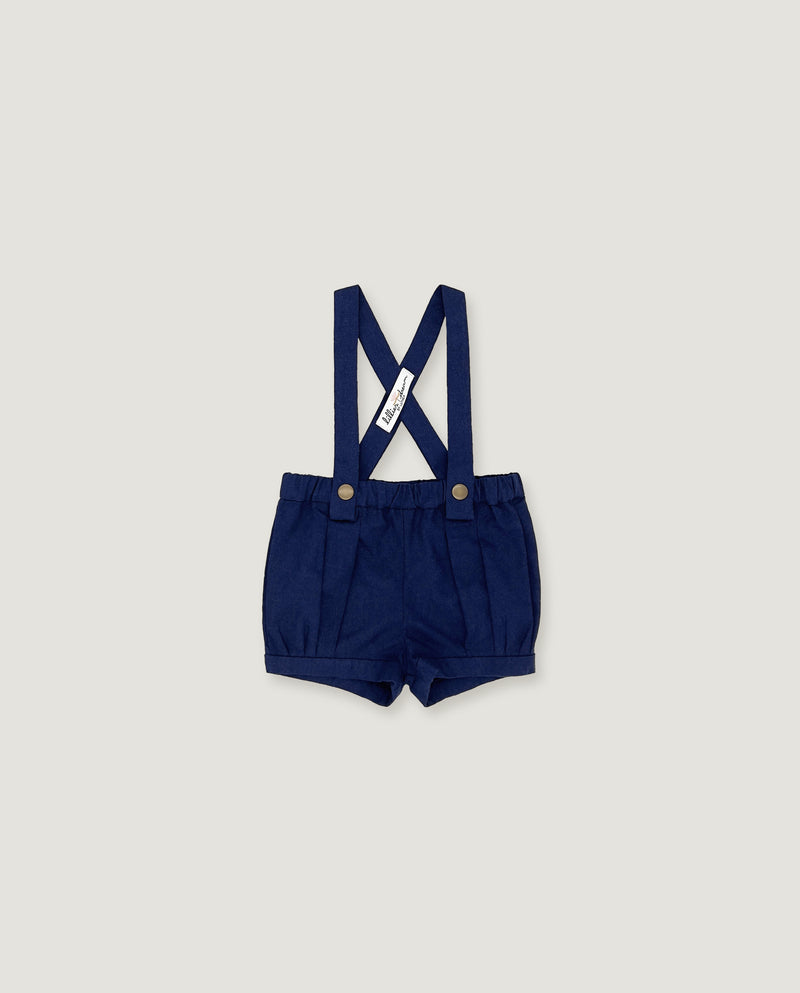 A pair of navy blue baby boy shorts with adjustable suspenders, featuring a white fabric brand tag and gold-tone snap buttons, displayed flat against a light gray background.