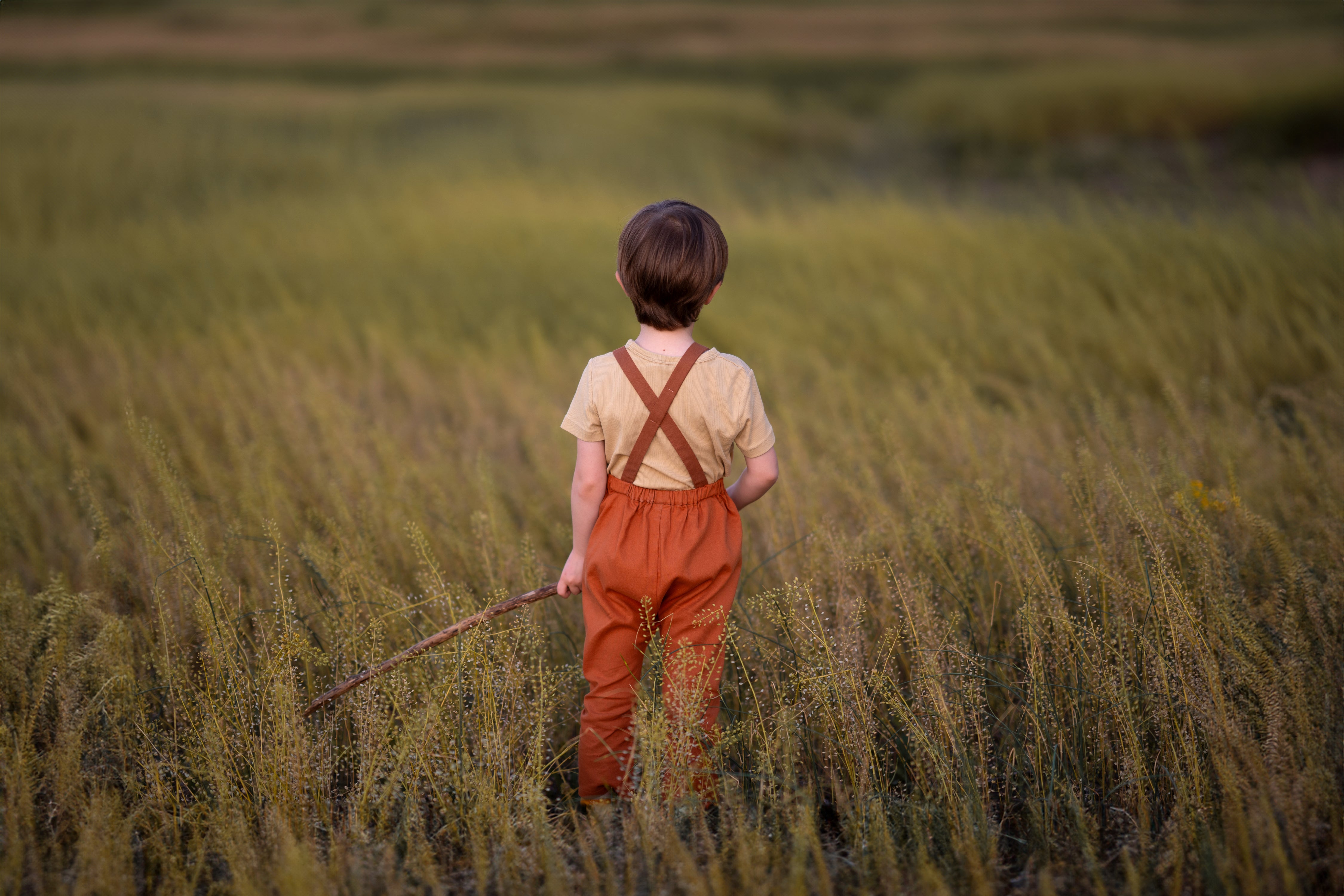 Child in orange overalls standing in a field holding a stick