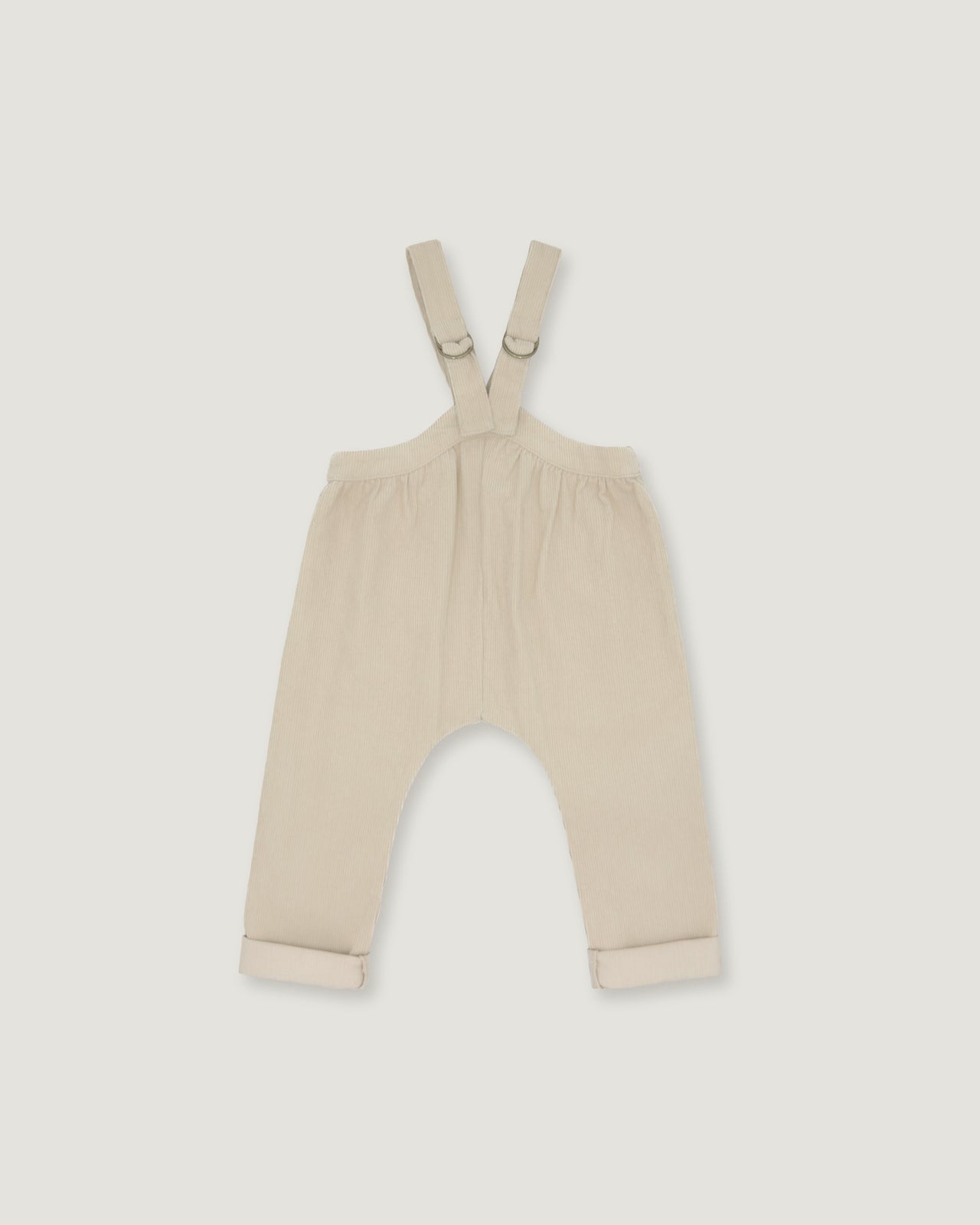 A pair of baby overalls in a light cream or beige color, featuring adjustable shoulder straps with metal rings and rolled cuffs at the bottom, shown flat against a light gray background.